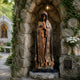 Monumental Traditional Virgin Mary Bronze Statue by Giant Sculptures in a stone grotto shrine with lantern, ivy, white roses, and candles.