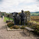 Life-Size Traditional Highland Bull and Herdsman Bronze Sculpture by Giant Sculptures displayed on heather-lined gravel path beside a stone wall and iron bench.