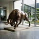 Giant Sculptures Life-Size Charging Bull bronze sculpture in lowered-head pose on a stone base in a modern lobby beside a glass-walled courtyard garden.