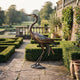 Traditional Standing Heron Bronze Sculpture by Giant Sculptures, 110cm, side view with arched neck, displayed on a stone terrace beside clipped box hedges.