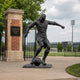 Giant Sculptures Life-Size Contemporary Football Player Bronze Sculpture 210cm shown mid-kick beside the Thompson Soccer Field gate with pitch behind.