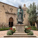 Monumental Traditional Our Lady of Guadalupe Bronze Statue, 400cm, teal mantle and gold-trimmed robe with sunburst halo, on stone base outside a church courtyard.