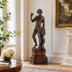 Classical Standing Bacchus Bronze Sculpture, 100cm, on a wooden pedestal in a living room, raising a chalice near a fireplace and framed art.