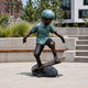Life-Size Contemporary Skateboarding Boy Bronze Sculpture, 130cm, arms spread with knees bent on a board atop a rock base, outdoor plaza setting.