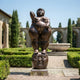 Monumental Rotund Nude Female Figure Bronze Sculpture, 350cm, standing on a large sculpted head, bronze finish, displayed on a garden stone path with hedges and arched colonnade behind.