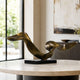 Bronze Abstract Flowing Ribbon Sculpture in brass on a black rectangular base, displayed on a round table in a living room near a window.