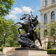 Saint George and the Dragon Bronze Statue - 350cm, showing the warrior on a rearing horse spearing the dragon, set outdoors near a stone building.