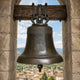 Large Classical Church Bell Bronze Sculpture, 120cm, cast bronze with relief panel, hung from a wooden beam in a stone archway overlooking a hillside town.