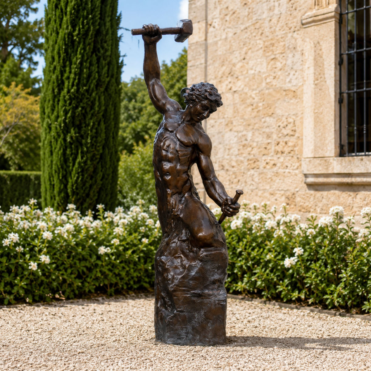 Large Classical Blacksmith Figure Bronze Sculpture, 250cm, muscular figure raising a hammer atop a rocky base, set on a gravel garden path near stone building and greenery.