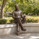 Life-Size Contemporary Seated Man and Cat Bronze Sculpture, 190cm, cast bronze figure seated on a granite bench beside a cat, outdoor garden setting with trees and flowering shrubs.