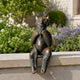 Traditional Seated Faun Bronze Sculpture, 80cm, cast bronze faun with horns and braided beard, hands to chin, seated on a stone ledge amid garden greenery.