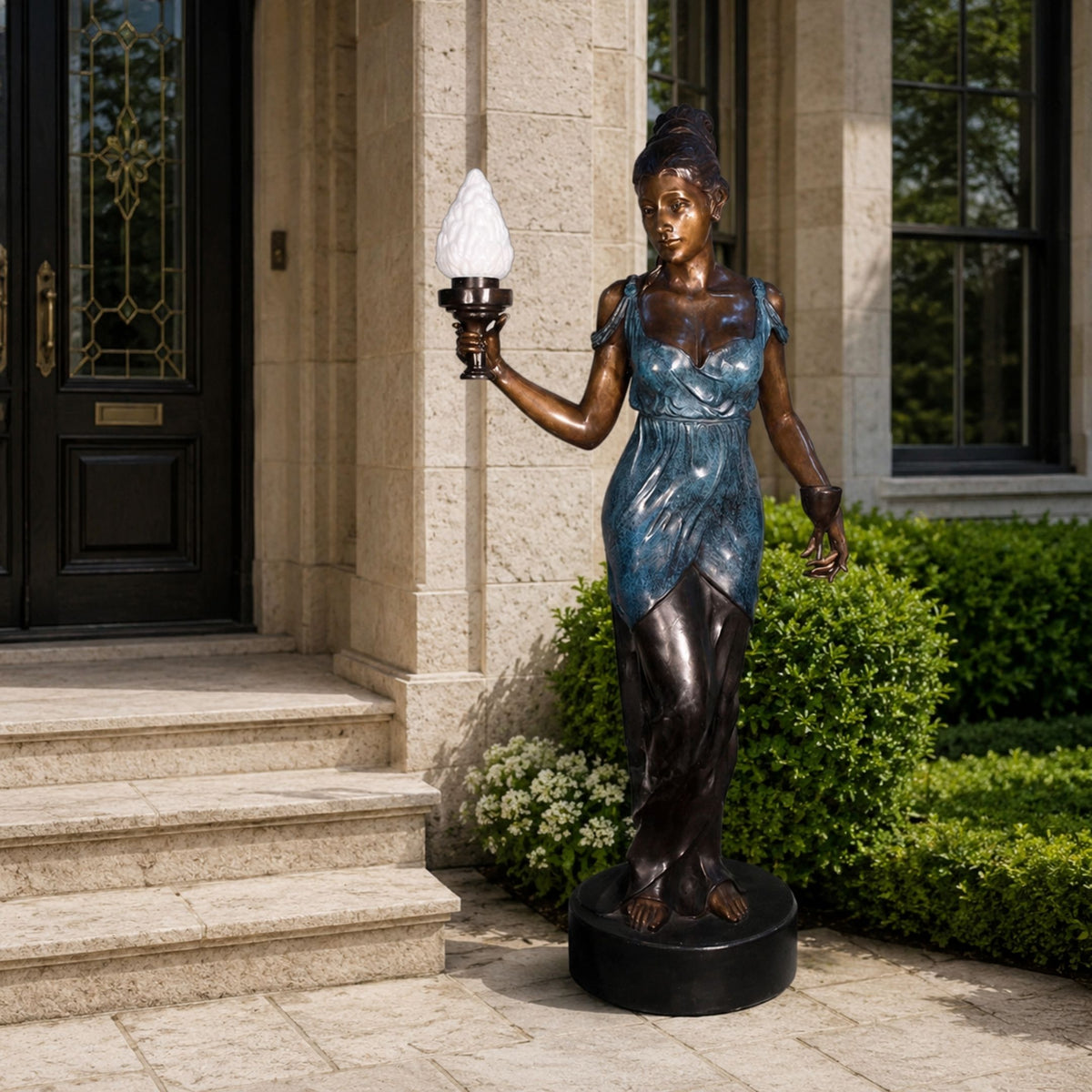 Large Classical Standing Female Figure Bronze Sculpture, 190cm, holding a torch lamp, on a stone base beside a home entrance with greenery.