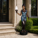 Large Classical Standing Female Figure Bronze Sculpture, 190cm, holding a torch lamp, on a stone base beside a home entrance with greenery.