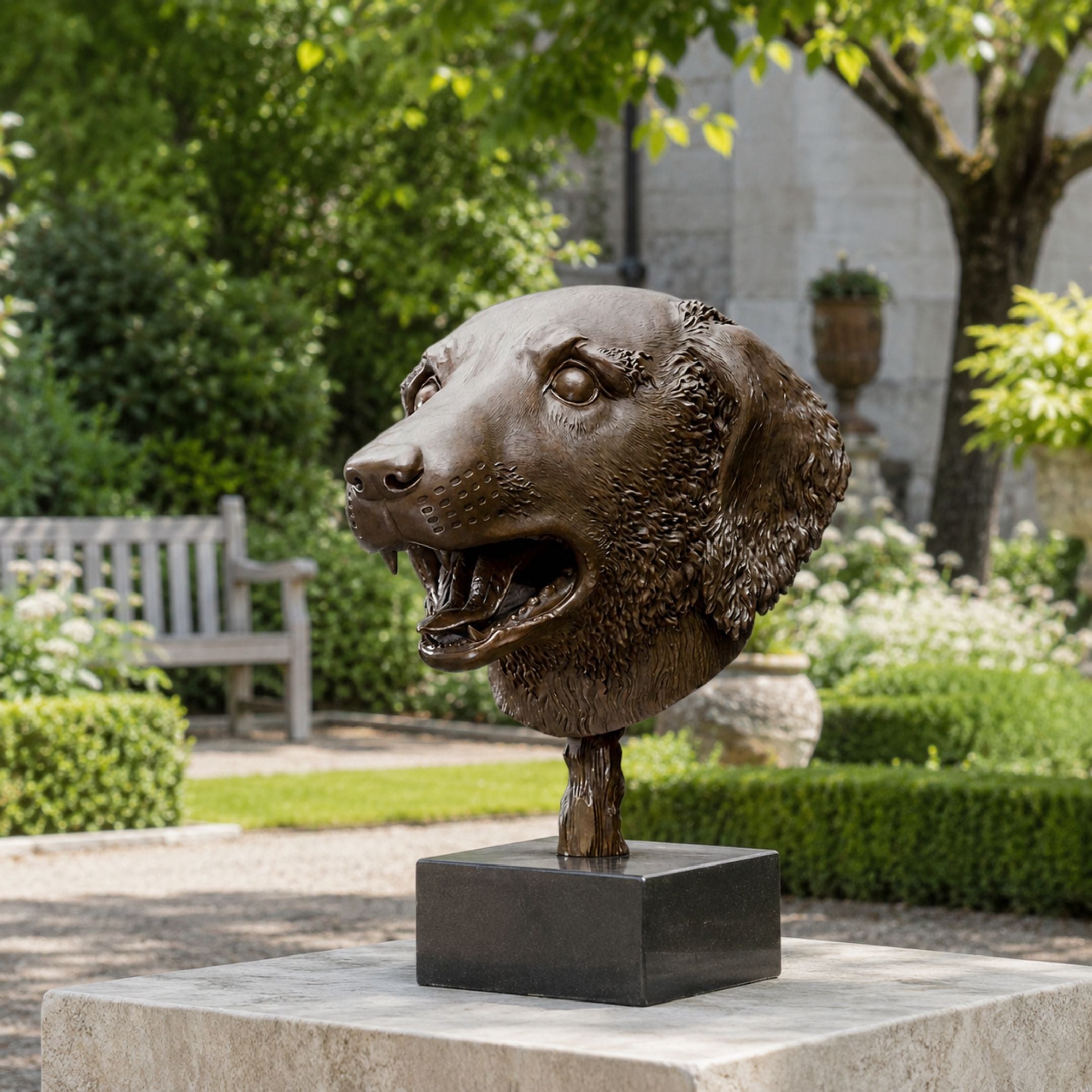 Contemporary Dog Head Bronze Sculpture, 45cm, on a dark stand atop a stone plinth in a garden setting with greenery and a bench nearby.