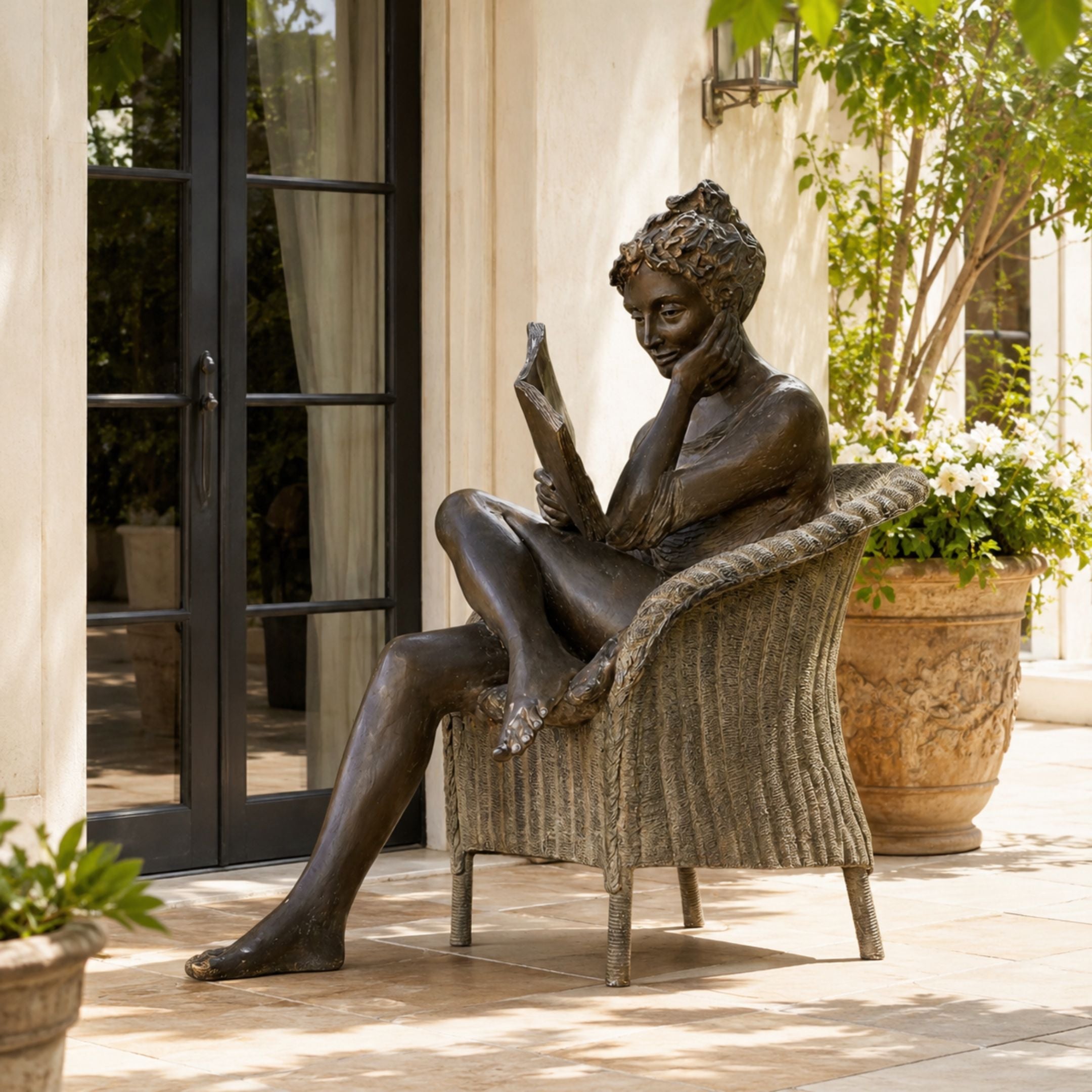 Contemporary Reading Woman Bronze Sculpture, 150cm, seated in a wicker-pattern armchair reading a book, patio setting near glass doors and potted plants.