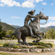 Life-Size Traditional Cowboy Riding Galloping Horse Bronze Sculpture, 400cm, by Giant Sculptures, set on a stone-edged garden bed with mountains and white fencing behind.