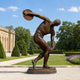 Life-Size Classical Discus Thrower Bronze Statue, 150cm, on a round base in a garden setting with hedges and a stone building behind.