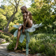 Life-Size Contemporary Seated Female Figure Bronze Sculpture, 250cm, nude figure with knees drawn up seated on a lotus-form perch atop a tall stem, set in a garden among greenery and flowers.