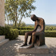Monumental Modern Seated Figure with Hollow Torso Bronze Sculpture, 220cm, bowed head and hollow torso, on a stone base outdoors amid hedges and trees.