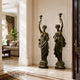 Large Classical Goddess Pair Bronze Sculpture - 180cm, two robed bronze figures raising torchères, displayed in an entryway beside a living room with beige sofa.