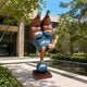 Giant Contemporary Rotund Figure Handstand Bronze Sculpture, 220cm, showing a rotund figure balancing inverted on a blue sphere with brown patina finish, displayed outdoors on a stone base near a reflecting pool.