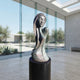 Modern Elongated Female Bust Bronze Sculpture, 150cm, on a black cylindrical base in a glass-walled room with a pool and desert plants visible beyond.