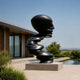 Monumental Modern Spiralling Head Bronze Sculpture, 250cm, black cast bronze face profile atop a twisting spiral, on a stone base beside a modern home and pool.