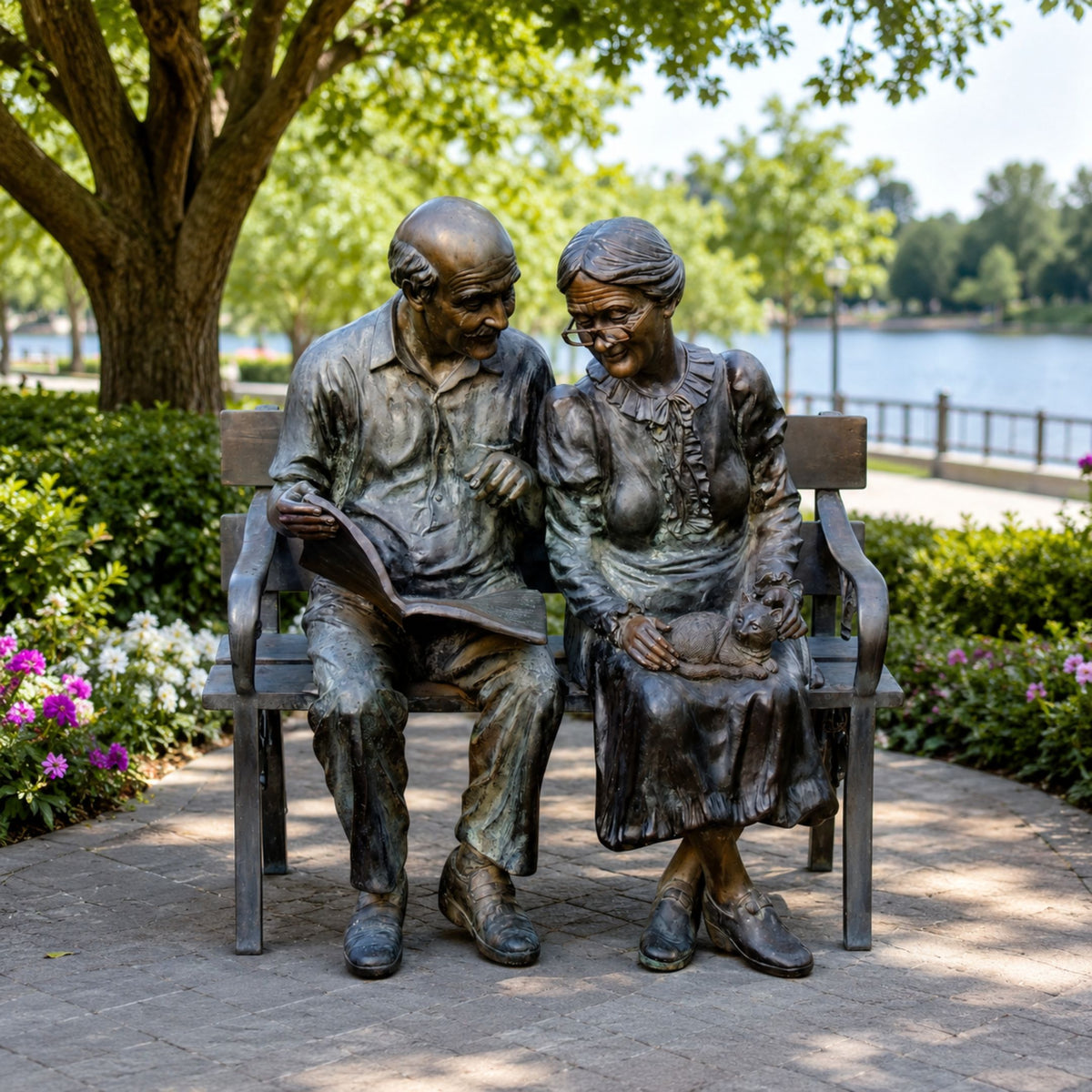 Life-Size Traditional Elderly Couple on Bench Bronze Sculpture (Book and Cat) - 170cm, shown outdoors on a park path with greenery and a lake in the background.