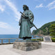 Monumental Traditional Samurai Warrior Bronze Statue, 350cm, on a stone base overlooking the coastline, with two swords at the waist and a weathered green-bronze finish.