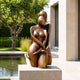 Modern Abstract Female Figure Bronze Sculpture, 100cm, on a stone plinth beside a reflecting pool with trees and a modern building facade behind it.