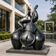 Monumental Modern Abstract Cellist Bronze Sculpture, 320cm, black patina figure playing a double bass, displayed outdoors on a stone plaza near a building and reflecting pool.