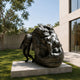 Large Contemporary Oversized Head and Figure Bronze Sculpture, 180cm, showing a giant tilted bronze head with a small nude figure leaning against it, set on a stone base in a courtyard.
