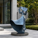 Large Contemporary Wind-Swept Female Figure Bronze Sculpture, 220cm, blue-toned patina on a round dark base, set on a courtyard patio near greenery and a modern building facade.