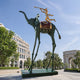 Monumental Contemporary Surrealist Elephant and Trumpeting Rider Bronze Sculpture, 600cm, with green-patina elephant on tall spindly legs and gilded trumpeter, installed on a plaza near a colonnaded building and arch.