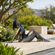 Life-Size Contemporary Reclining Woman Bronze Sculpture, 80cm, in bronze with wide-brimmed hat, reclining poolside on a stone patio amid greenery.