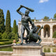 Monumental Classical Hero and Dragon Bronze Sculpture, 250cm, showing the nude hero raising a club over a rearing dragon, set on a stone pedestal in a garden courtyard.
