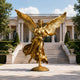 Monumental Classical Winged Angel Bronze Sculpture in gilded gold, 300cm, spread eagle-style wings and windswept robes, displayed on a round base before a colonnaded building.