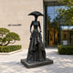 Monumental Contemporary Elongated Female Figure Bronze Sculpture, 350cm, dark bronze finish with wide-brimmed hat and layered gown, on a stone plaza outside an art gallery entrance.