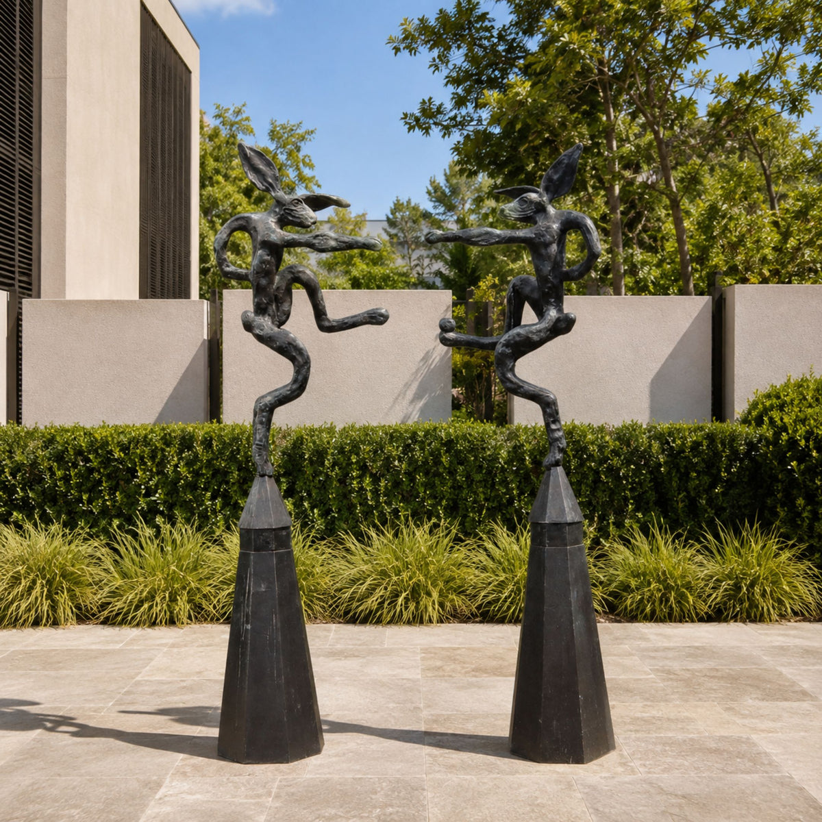 Large Contemporary Fighting Hares Bronze Sculpture, 180cm, two black bronze hare figures boxing atop tapered plinths in a landscaped courtyard.