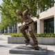 Life-Size Skateboarding Figure Bronze Sculpture, 250cm, showing a bronze male figure mid-ride with arms outstretched, displayed on a stone plinth outdoors near trees and a building.