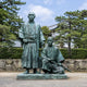Monumental Traditional Standing and Seated Samurai Bronze Sculpture (350cm) by Giant Sculptures, green-patina bronze figures in robes outdoors on stone base near trees and a traditional building.