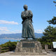 Monumental Traditional Samurai Warrior Bronze Sculpture - 350cm by Giant Sculptures, arms folded and katana at hip, standing on a stone pedestal overlooking a coastal mountain landscape.