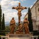 Monumental Classical Crucifixion Group Bronze Sculpture, 600cm, showing Christ on a cross with three robed mourning figures, set outdoors near stone buildings and trees.