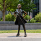Life-Size Contemporary Defiant Girl Bronze Sculpture, 130cm, hands on hips, gazing upward, cast bronze finish, displayed outdoors on a stone platform near greenery.