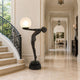 Life-Size Contemporary Nude Female Figure Bronze Sculpture, 210cm, in dark bronze finish leaning forward on tiptoe to hold a glowing globe lamp, displayed at the base of a staircase in a marble hallway.