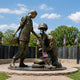 Monumental Contemporary Female Soldier Memorial Bronze Sculpture, 350cm, showing two bronze female soldiers at a battlefield cross with rifle, helmet, and boots, set outdoors near a memorial wall.