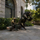 Life-Size Contemporary Bull and Man Reading Bronze Sculpture by Giant Sculptures, dark rearing bull and golden-brown seated man sharing a bench outside a stone building.