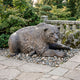 Large Contemporary Bear with Fish Bronze Sculpture by Giant Sculptures, shown crouched on river pebbles in a garden setting beside a stone balustrade and maples.