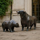 Bronze bear and bull sculptures by Giant Sculptures facing each other on stone paving outside a classical stone building with ivy and tall windows.
