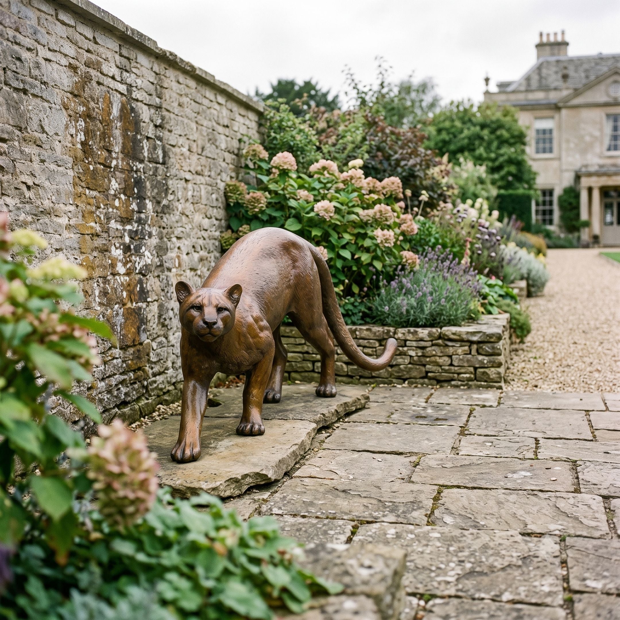 Life-Size Traditional Stalking Cougar Bronze Sculpture by Giant Sculptures in warm golden-brown patina, positioned on a stone garden path beside hydrangeas and a manor house.